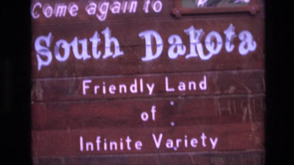Close 1974 8mm view of the South Dakota welcome sign, emphasizing the painted white lettering and the phrase Friendly Land of Infinite Variety.