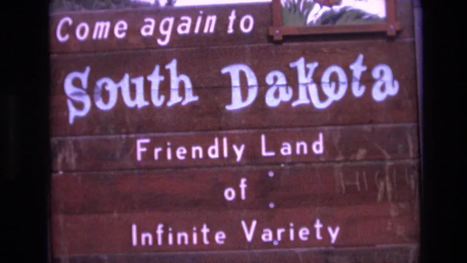 Tight 8mm frame of the South Dakota sign, showing large painted state-name lettering across weathered wood and the lower tourism slogan text.