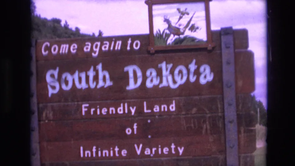 Angled 1974 8mm view of the South Dakota welcome sign, with the wooden structure, white painted message, decorative panel, and surrounding roadside vegetation visible.