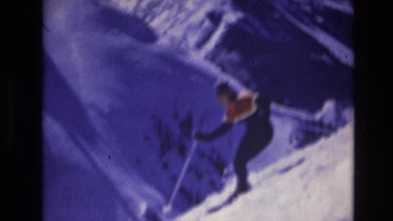 A skier in dark clothing and a bright orange vest descends a snowy Vail, Colorado slope in soft blue-tinted 1974 8mm footage.