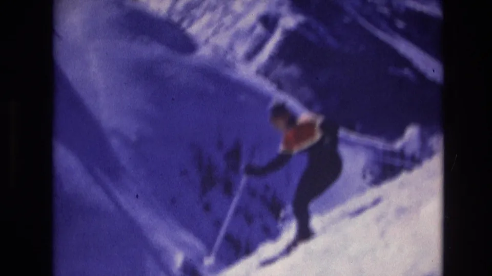 A skier in dark clothing and a bright orange vest descends a snowy Vail, Colorado slope in soft blue-tinted 1974 8mm footage.