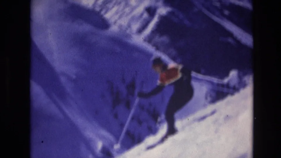 A skier in orange and dark clothing continues down the snowy mountain slope, captured with soft focus and handheld 8mm color texture.