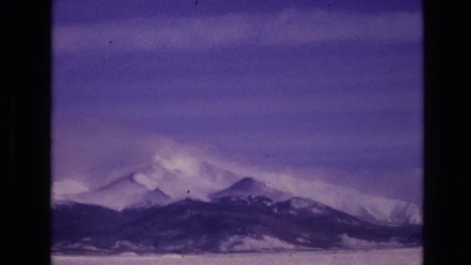 1974 Vail, Colorado 8mm frame with a wide alpine view of snow-covered peaks and dark lower ridges beneath a cloudy sky.