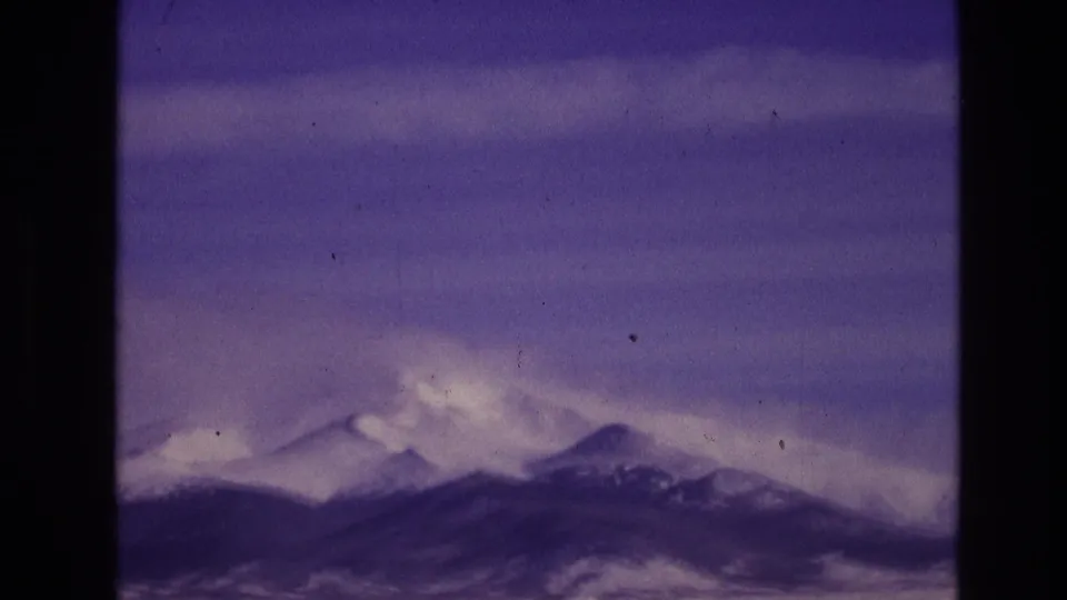 1974 8mm home-movie view near Vail, Colorado showing soft snowy peaks and a darker ridge beneath a broad purple-blue sky with visible film texture.