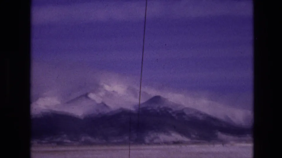1974 Vail, Colorado 8mm frame of distant snow-covered peaks and a dark mountain ridge, tinted purple with visible scratches, dust, and home-movie softness.