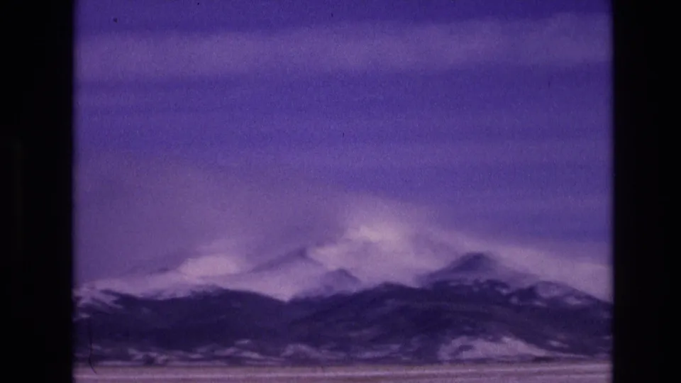 1974 Vail, Colorado home-movie frame showing a soft purple-toned mountain skyline with snow on the summits, a flat foreground, and visible 8mm film grain.