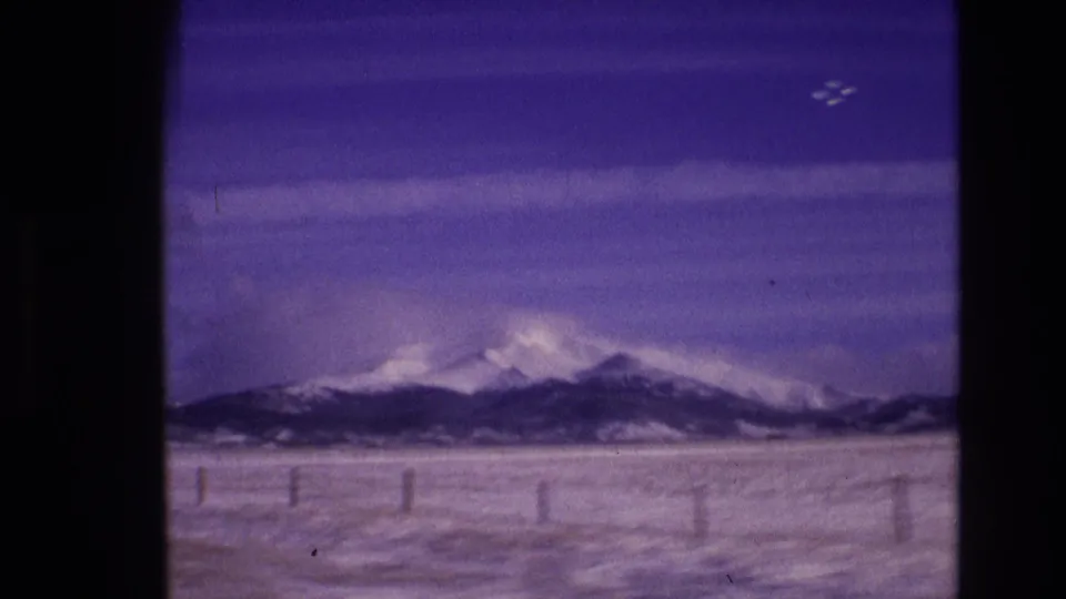 A moving-car view across a snow-blanketed plain toward Colorado mountain ridges, with faint fence posts and visible 8mm grain and color shift.
