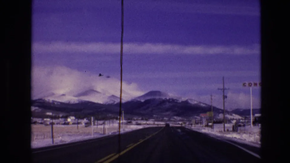 1974 Vail, Colorado road-view frame from a moving car, showing a dark two-lane road, snowy shoulders, utility poles, distant buildings, mountains, and partial roadside lettering.
