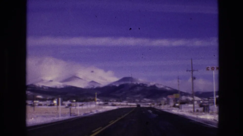 1974 8mm home-movie frame from Vail, Colorado, showing a dark road, snow-covered roadside, utility poles, and a distant mountain range.