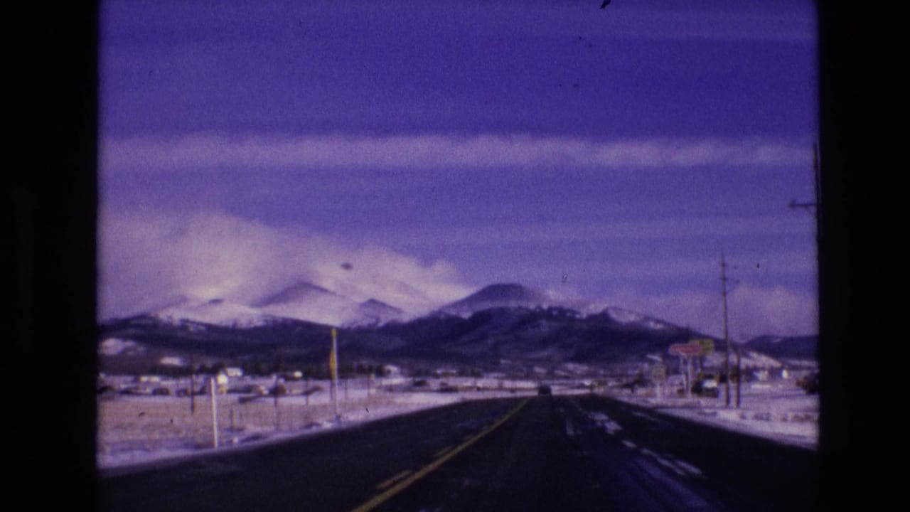 Straight road receding toward snow-covered mountains with poles and roadside structures.