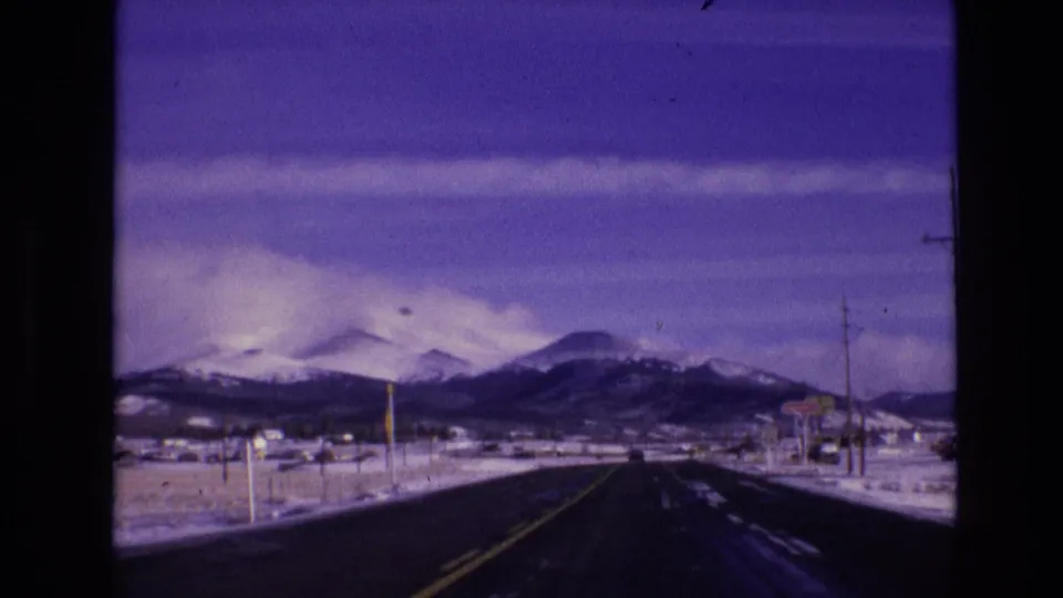 Vail, Colorado winter road scene from 1974 8mm footage, with the road stretching into a snowy mountain range and small roadside features along the route.