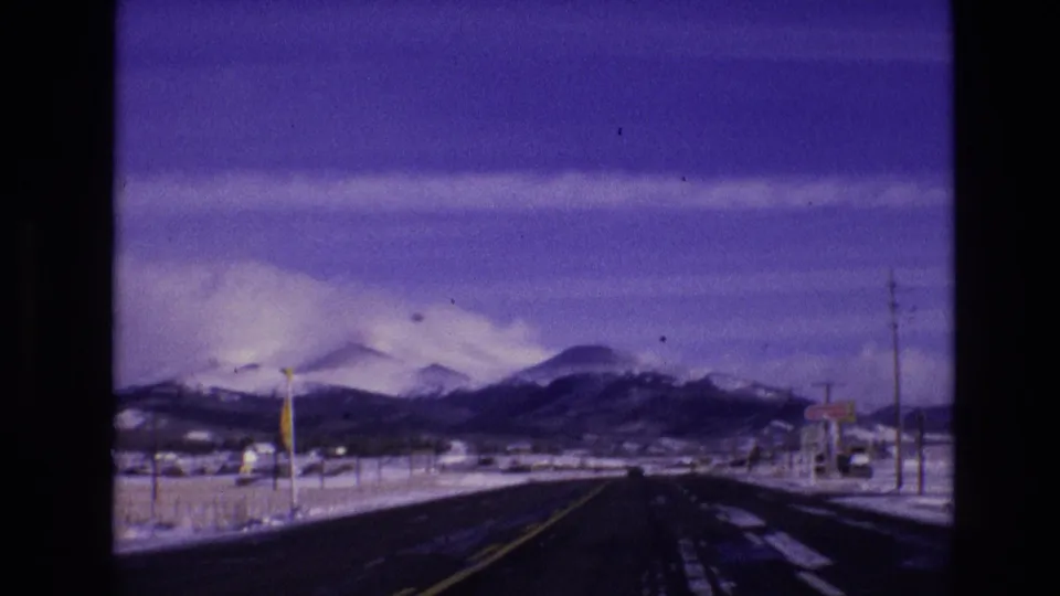 A 1974 Vail, Colorado travel frame showing a paved road from a moving vehicle, with snowbanks, roadside poles, small signs, and mountains ahead.