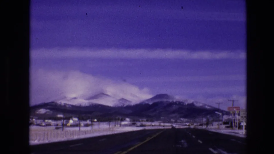 Final frame of the Vail, Colorado road approach, with a long dark roadway, snow-covered shoulders, poles, distant settlement details, and the mountain range beyond.