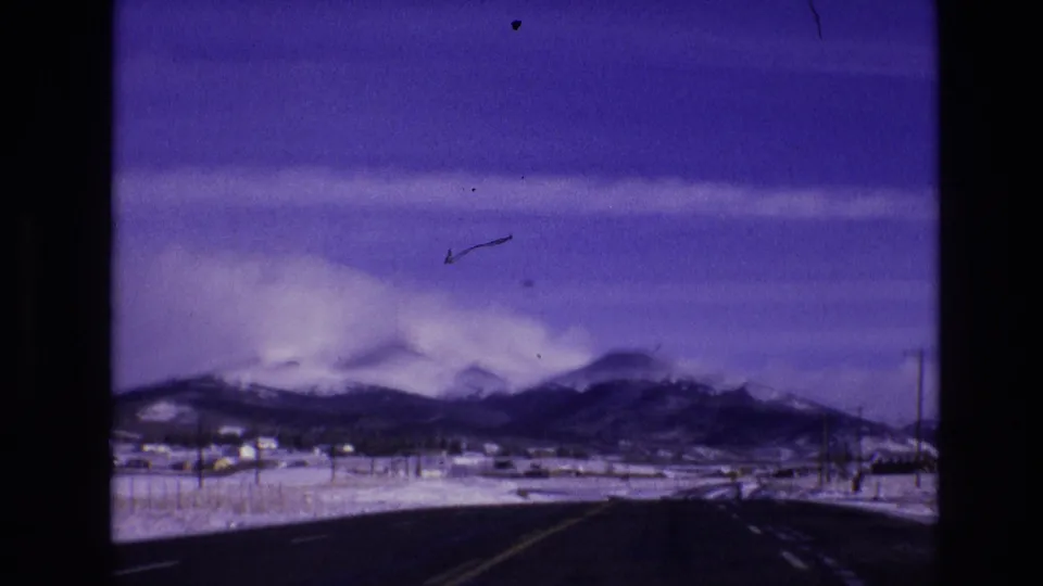 1974 Vail, Colorado 8mm frame showing a moving-vehicle view of a snowy roadside valley and mountain range with visible film debris or a passing mark near the sky.