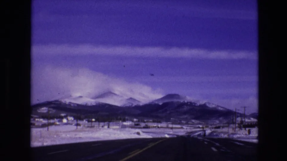 1974 Vail, Colorado home-movie frame showing a winding road through snowy ground toward dark mountain ridges and a clouded purple sky.