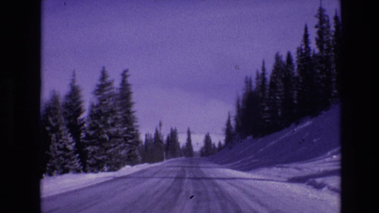 1974 Vail, Colorado home-movie frame showing a snow-covered road ahead through evergreen forest under a purple-blue winter sky.