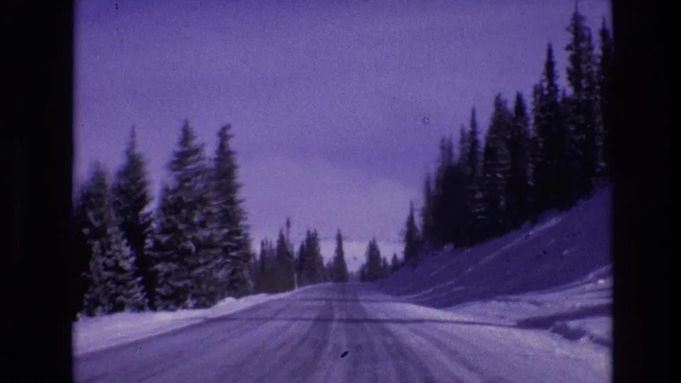 1974 Vail, Colorado home-movie frame showing a snow-covered road ahead through evergreen forest under a purple-blue winter sky.