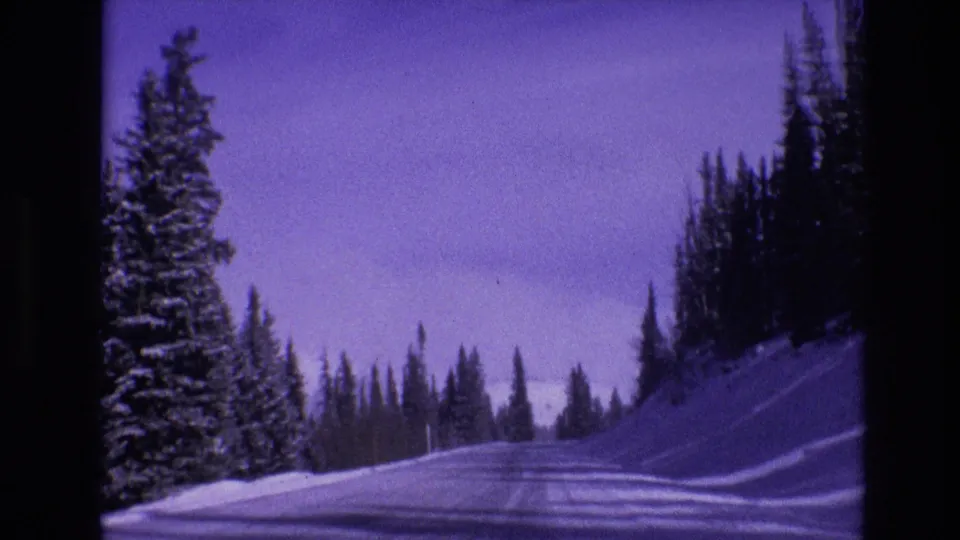 1974 Vail, Colorado 8mm frame showing a snow-covered road receding between evergreen trees, snowbanks, and distant mountain terrain.