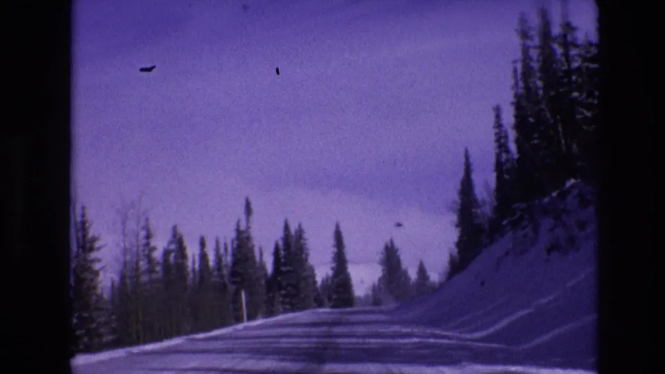 1974 Vail, Colorado 8mm frame showing a snow-covered road running past dark evergreens, with tire tracks, snowbanks, and visible film dust.