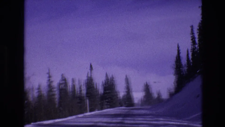 1974 Vail, Colorado 8mm frame with a soft forward view of a snowy mountain road, evergreen trees, tire tracks, and a strong purple archival color cast.