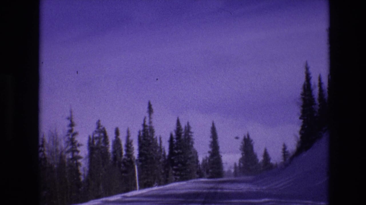 1974 8mm home-movie frame from Vail, Colorado showing a snowy roadside, tall evergreen trees, and a blue-purple mountain sky from a moving vehicle perspective.