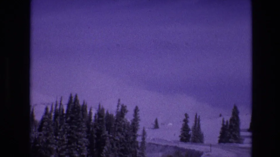 1974 Vail, Colorado 8mm frame with a curving snowy mountain road, clustered evergreens, distant slope, and a small dark speck in the sky area.