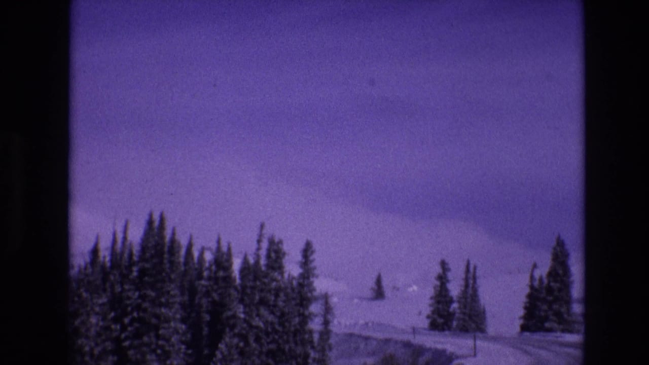 1974 Vail, Colorado home-movie frame showing a snow-covered highway bending through evergreen trees with distant white slopes and aged-film purple color shift.