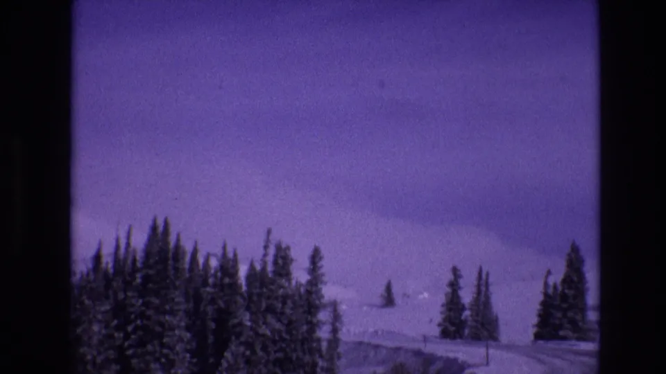 1974 Vail, Colorado home-movie frame showing a snow-covered highway bending through evergreen trees with distant white slopes and aged-film purple color shift.