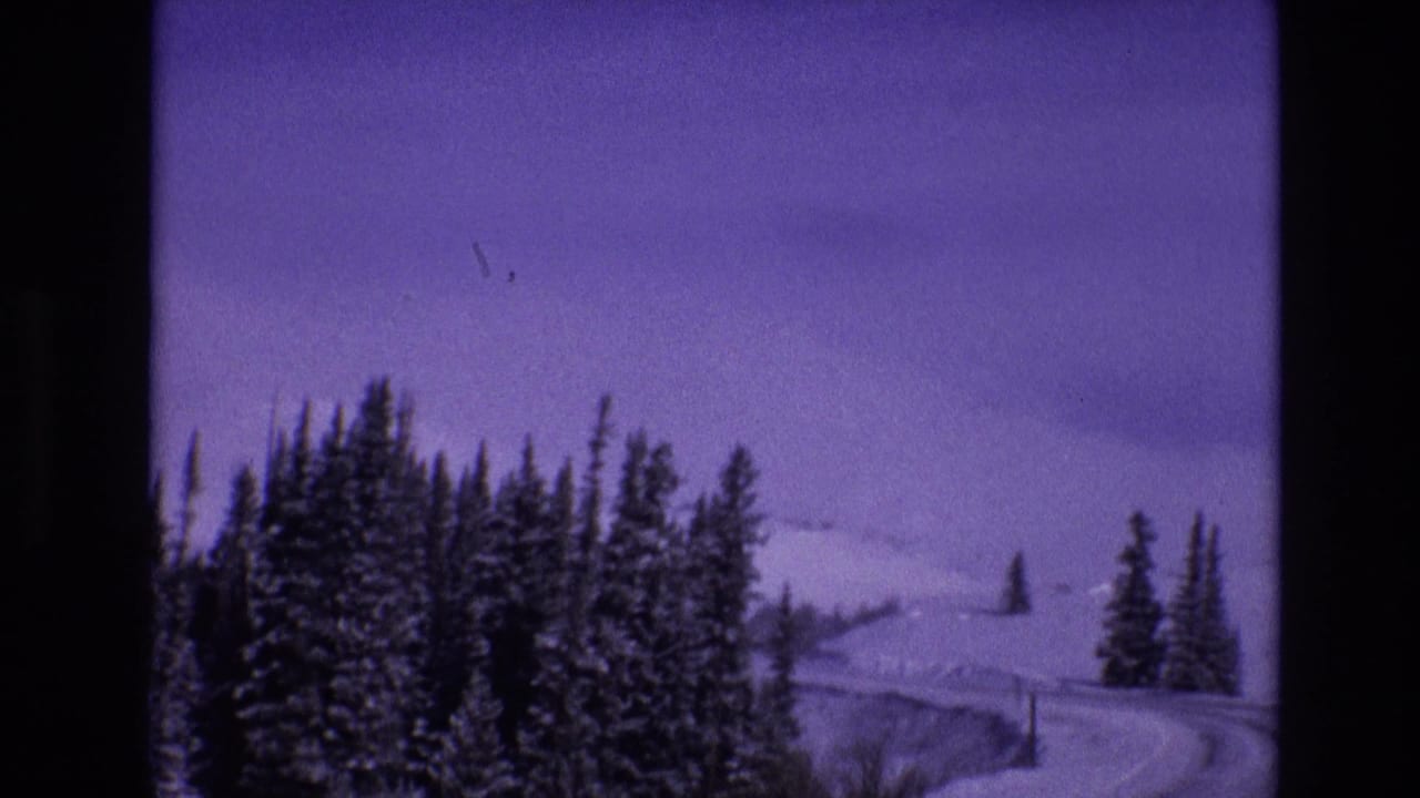 1974 Vail, Colorado frame with dense snow-dusted evergreens, a winding mountain highway, roadside poles, and pale snowy slopes in the background.