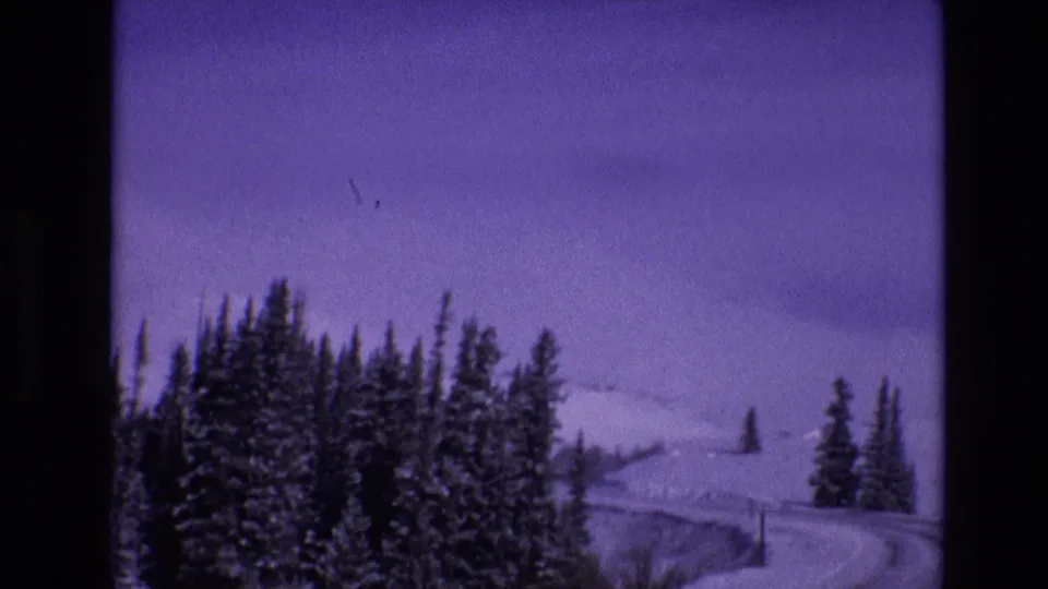 1974 Vail, Colorado frame with dense snow-dusted evergreens, a winding mountain highway, roadside poles, and pale snowy slopes in the background.