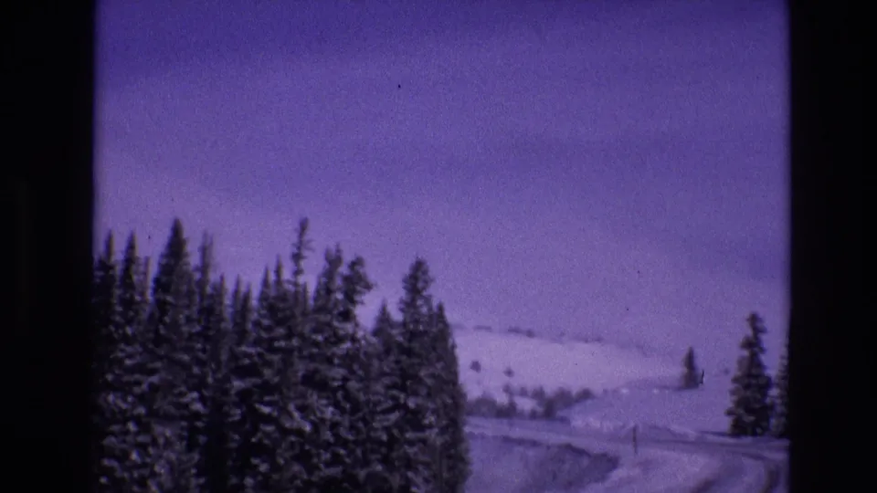 1974 Vail, Colorado home-movie frame showing roadside evergreens, a snow-covered curve, and an open white hillside seen from a moving vehicle.