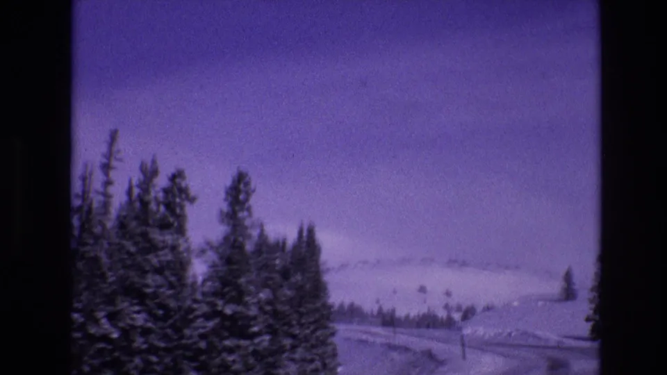 1974 Vail, Colorado frame with dark evergreen trees in the foreground, a snowy highway edge, roadside poles, and distant mountain snowfields under a purple-tinted sky.
