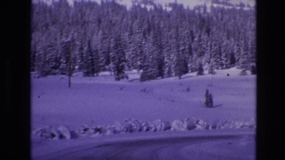 1974 Vail, Colorado 8mm frame showing a broad snowy foreground, plowed roadside edge, dense evergreen forest, and faint frozen surface near the lower frame.