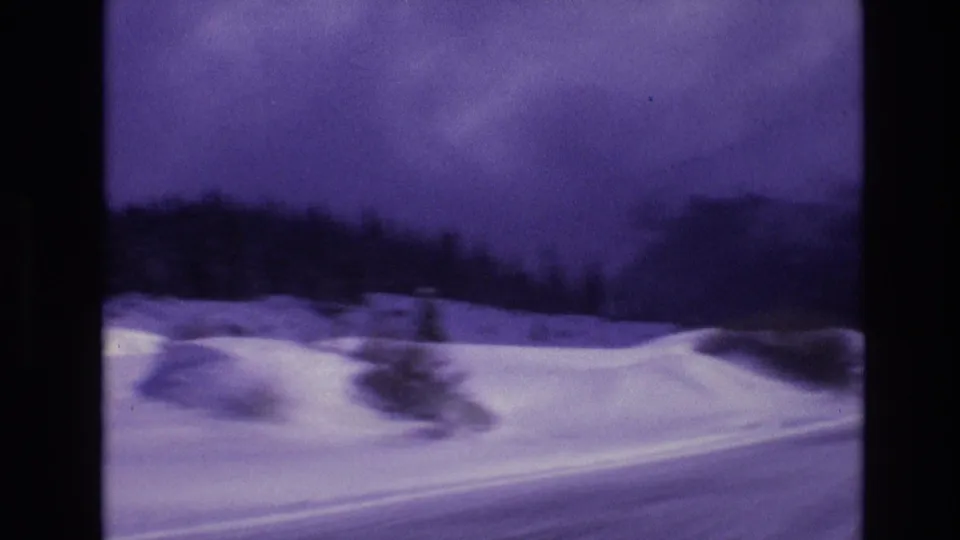 1974 Vail, Colorado 8mm frame showing a snowy roadside foreground, dark evergreen tree line, and purple-blue winter sky seen from a moving vehicle.