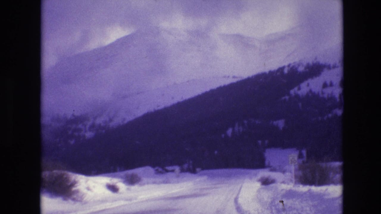 Snowy road curves through a mountain landscape with a roadside sign and small structure.