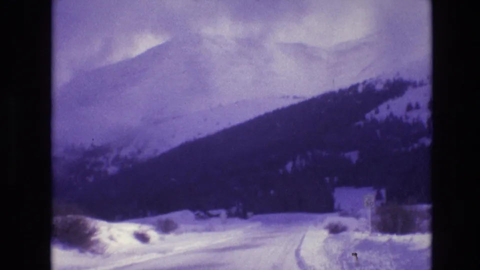 1974 Vail, Colorado home-movie frame showing a snow-covered road, mountain slopes, a roadside speed-limit sign, and a small structure partly obscured by snow and blur.