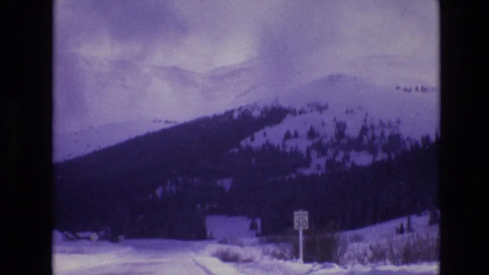 1974 Vail, Colorado 8mm frame with a dark mountain slope, snowy roadside, distant pale ridge, and a visible speed-limit sign near the road edge.