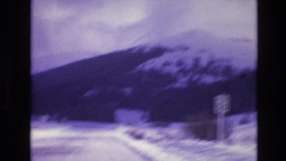 1974 Vail, Colorado frame showing a motion-blurred winter road view with snowbanks, a roadside sign, a small structure, and dark mountain slopes beyond.