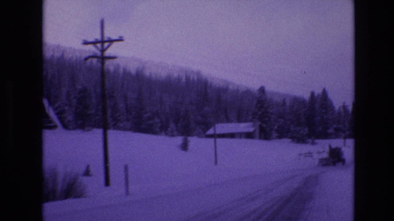 1974 Vail, Colorado 8mm frame showing a snow-covered road beside utility poles, a small cabin, evergreen trees, and a distant snowplow in a purple-tinted winter landscape.