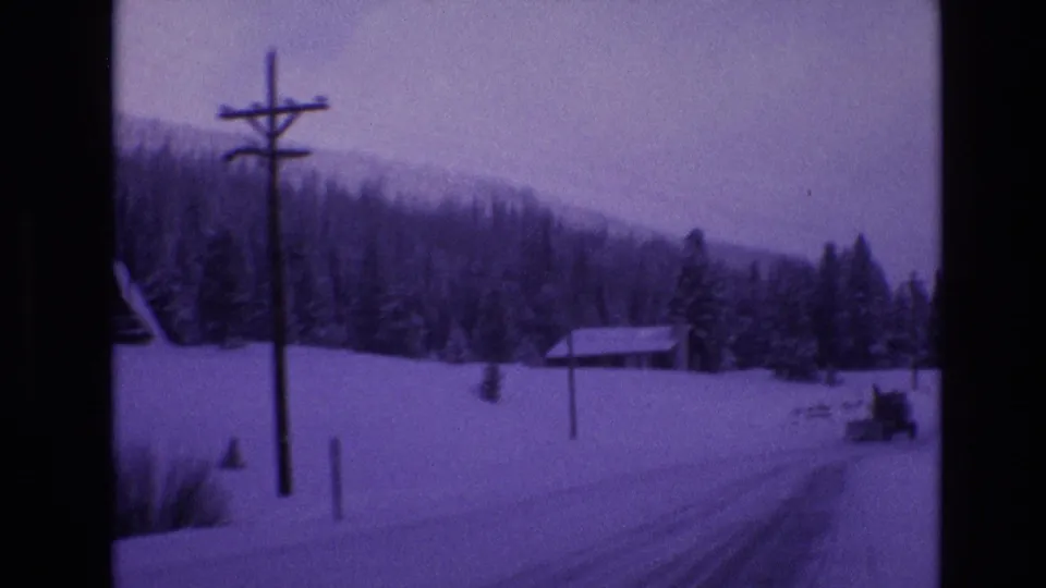 1974 Vail, Colorado 8mm frame showing a snow-covered road beside utility poles, a small cabin, evergreen trees, and a distant snowplow in a purple-tinted winter landscape.