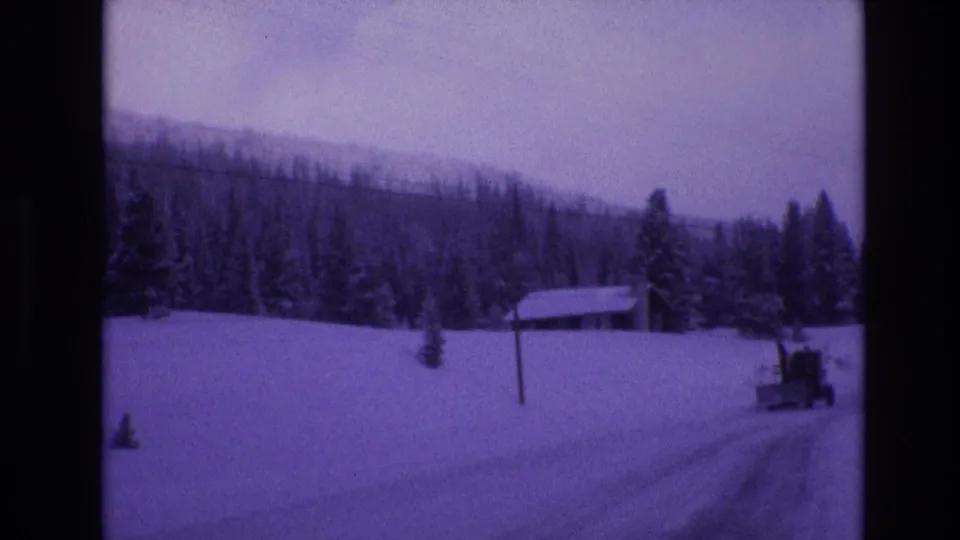 1974 Vail, Colorado frame with a snowplow moving along a snow-covered rural road near a small cabin, utility pole, evergreens, and wooded mountain slope.