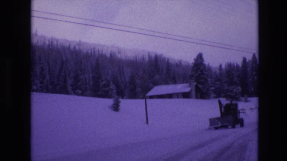 1974 Vail, Colorado 8mm frame showing a blade-front snowplow on a snowy road below overhead utility wires, with a cabin and forested hillside in the background.