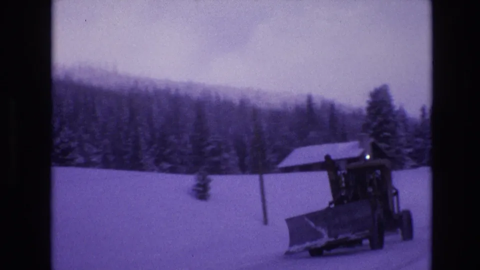 1974 Vail, Colorado frame showing a closer view of a snowplow and front blade on the winter road, with a small cabin, evergreen trees, and wooded slope behind it.