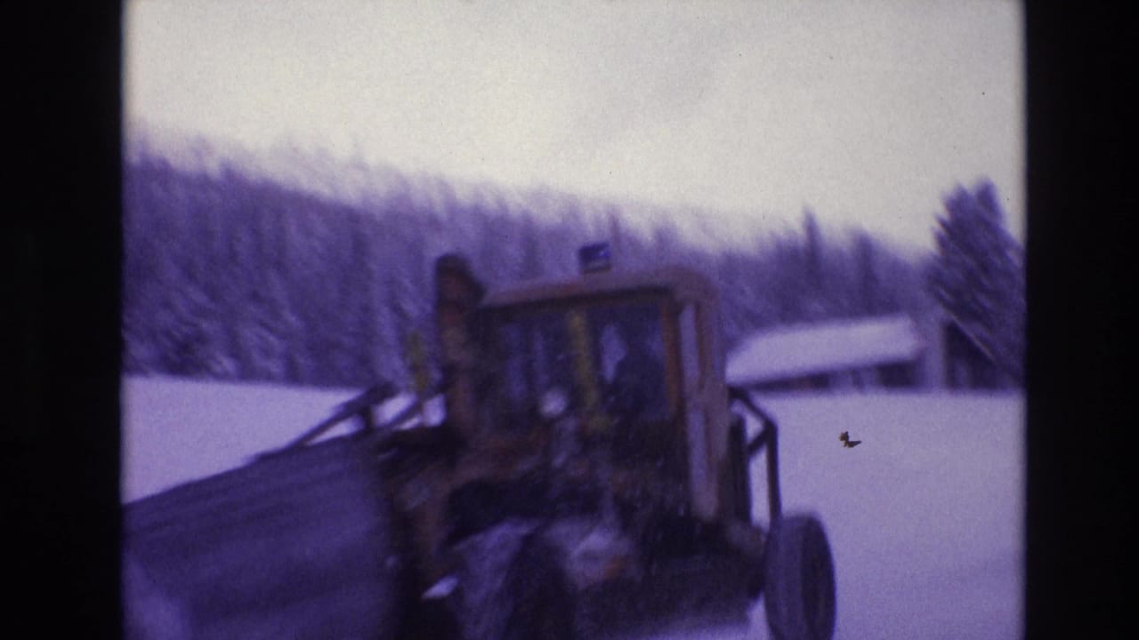 1974 Vail, Colorado home-movie frame showing a tractor in deep snow with a small snow-covered structure and wooded hillside behind it.