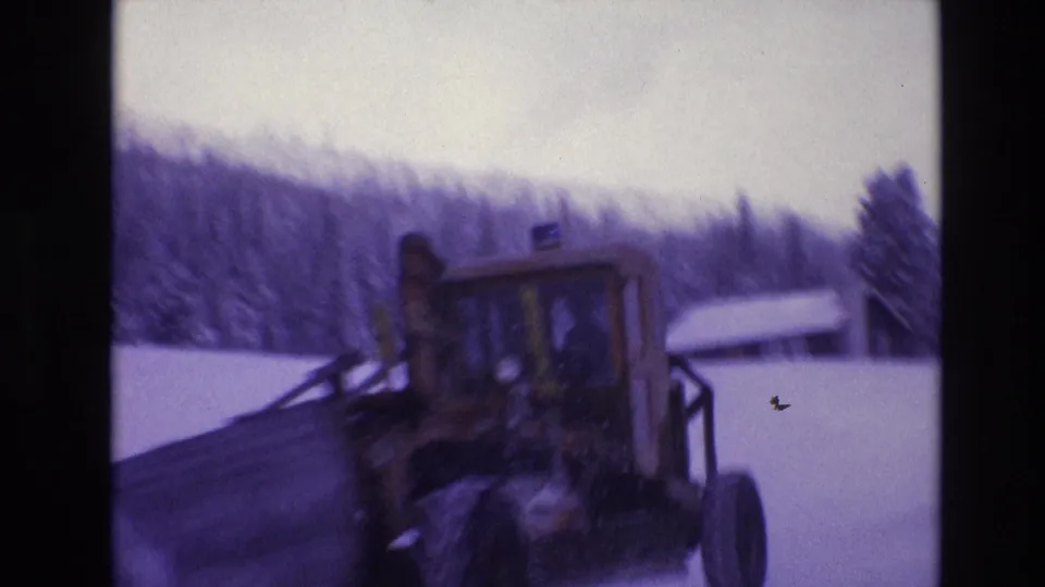 1974 Vail, Colorado home-movie frame showing a tractor in deep snow with a small snow-covered structure and wooded hillside behind it.