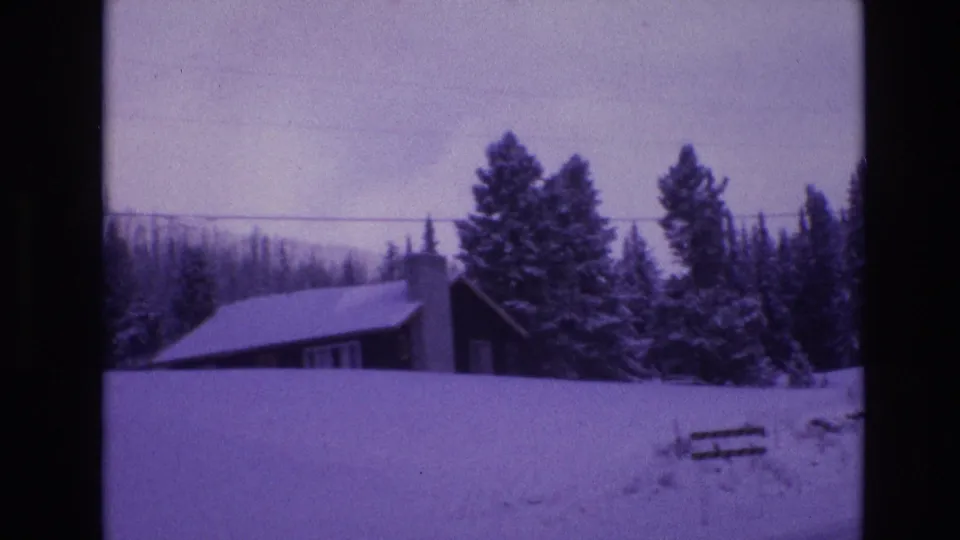 1974 Vail, Colorado frame showing a small cabin roofline and chimney behind a snowbank, with evergreen trees and a fence section nearby.
