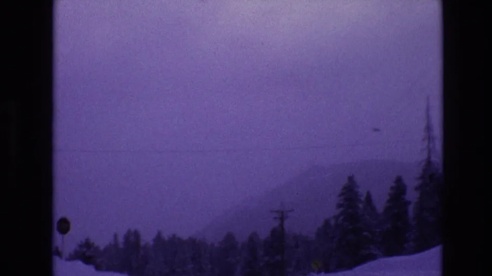 1974 Vail, Colorado home-movie frame showing a snow-covered roadside with utility poles, clustered evergreens, and a misty mountain horizon.