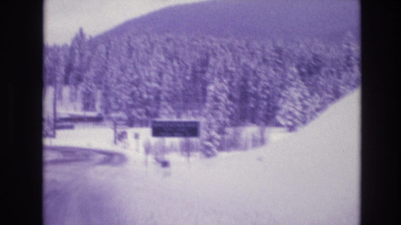 Purple-tinted 1974 Colorado 8mm frame showing a snowy roadside approach, evergreens, fencing, distant mountain slope, and an unreadable dark sign ahead.