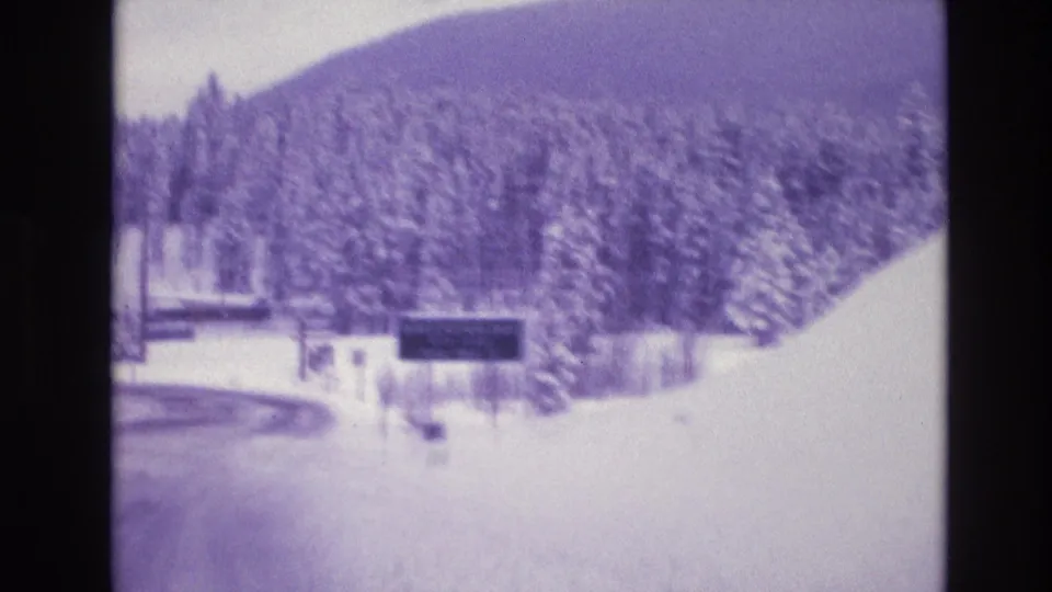 Purple-tinted 1974 Colorado 8mm frame showing a snowy roadside approach, evergreens, fencing, distant mountain slope, and an unreadable dark sign ahead.