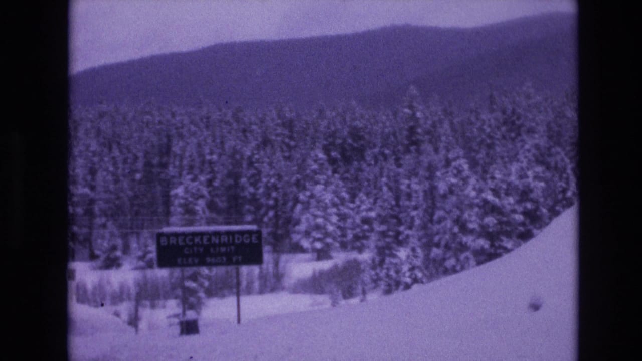 Purple-shifted 8mm frame of a Breckenridge city limit sign beside deep snow, evergreen trees, and a low mountain ridge in Colorado.